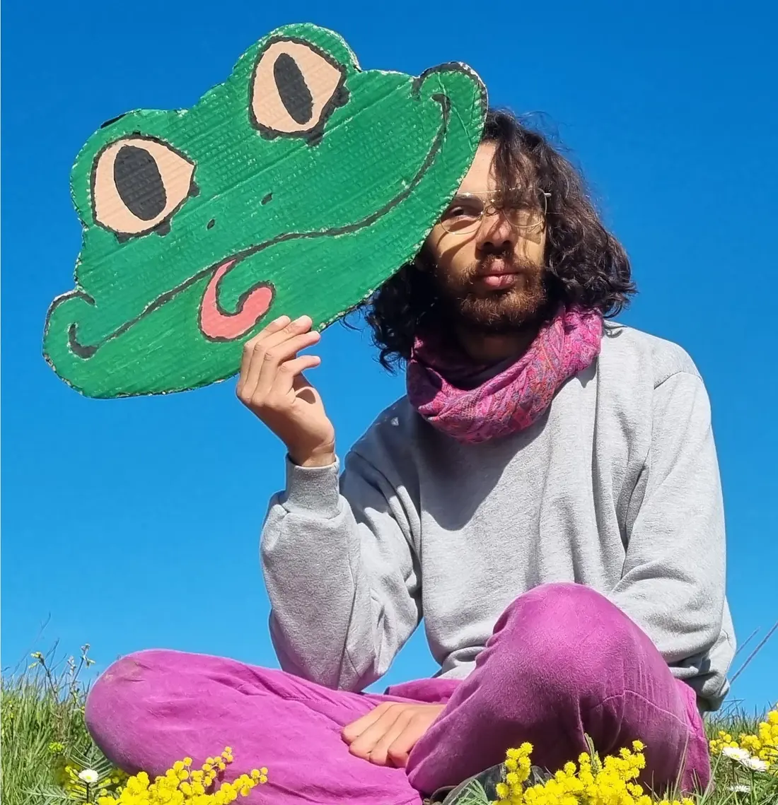 Vie, assis·e dans un champ de fleurs, tenant un masque de grenouille en carton